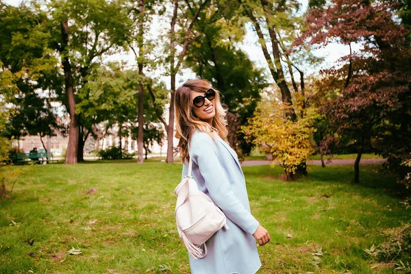 秋の公園でポーズをとるスタイリッシュなピンクのバックパックで青いコートとサングラスを着て若い幸せな女の屋外のポートレート — ストック写真