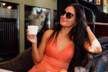 mutlu gülümseyen kadın güneş gözlüğü takıyor ve kahve fincanı tutarak açık kafe masada oturan