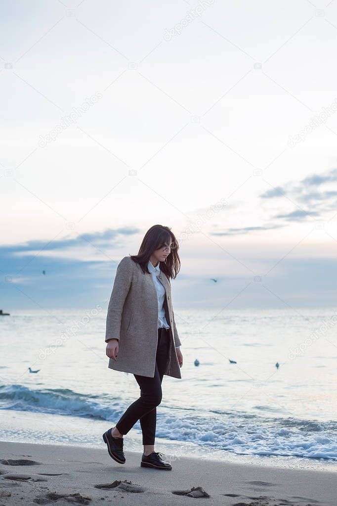 full length portrait of stylish brunette woman with grey coat walking on sandy beach