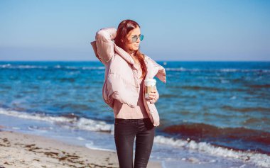 Kahve tutarak ve kumsalda yürüyen genç bir kadın