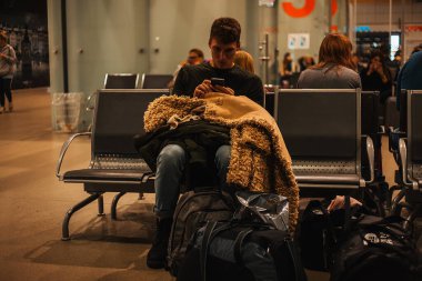 Uluslararası havaalanı terminalinin kalkış salonunda bavulla otururken akıllı telefon kullanan bir adam.