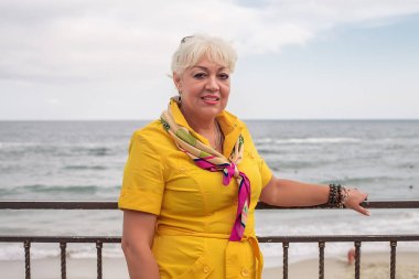 Deniz kıyısında poz veren sarı elbiseli, saç stili ve makyajlı güzel yetişkin bir kadın. 