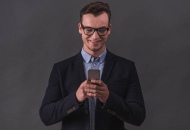 Takım elbise ve gözlük yakışıklı genç işadamı bir akıllı telefon kullanarak ve gri arka plan üzerinde gülümseyen