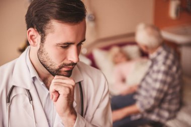 Yaşlı adam ön planda yakışıklı doktor hastane koğuşunda yatakta yatan karısını ziyareti yapıyor