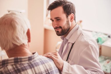 Yakışıklı Doktor yaşlı bir adamla konuşmak ve hastane koğuşunda otururken gülümseyen