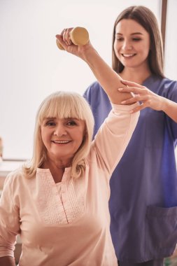 Güzel yaşlı kadın çalışmaları ile halter, kameraya bakarak ve, gülümseyen hastane koğuşunda yapıyor. Çekici hemşire ona yardım ediyor