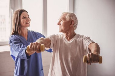 Yakışıklı ihtiyar dumbbells ile yapıyor ve, gülümseyen hastane koğuşunda. Çekici hemşire ona yardım ediyor