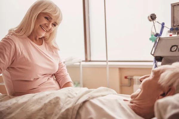 Vrolijke Oude Vrouw Kijkt Haar Man Liggend Bed Het Ziekenhuis — Stockfoto