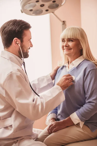 ハンサムな医者は 聴診器を使用して老婦人のハートビートを聴くこと 両方が笑っています — ストック写真