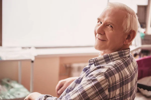 Handsome old man is looking at camera and smiling while sitting in wheelchair in hospital ward