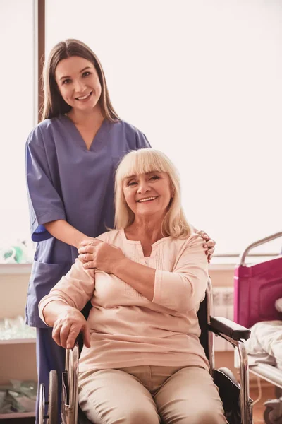 美丽的老太太正在看着相机 坐在轮椅上 在医院病房里微笑着 有魅力的护士就在附近 — 图库照片