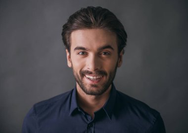Yakışıklı emin işadamı kıl, koyu mavi gömlek ile kameraya bakarak ve, gri arka plan üzerinde gülümseyen