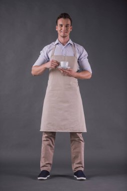 Tam uzunlukta bir holding, kameraya bakıyor ve gülümsüyor, gri arka plan üzerinde önlük yakışıklı genç barista görüntüsünü
