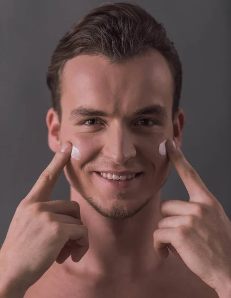 Hombre Joven Guapo Está Aplicando Crema Facial Mirando Cámara Sonriendo — Foto de Stock