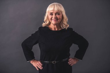 Şık elbiseli güzel eski Bayan kameraya bakarak ve gri arka plan üzerinde akimbo ayakta iken gülümseyen