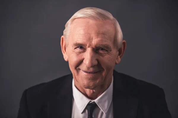 Handsome old businessman in suit is looking at camera and smiling, on gray background