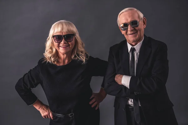 Beautiful old couple in elegant clothes and glasses is holding hands looking at camera and smiling, on gray background