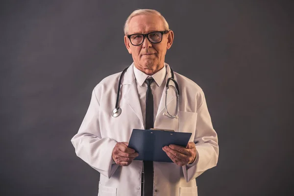 英俊的老医生在医疗大衣和眼镜持有一个文件夹 看着相机 在灰色背景 — 图库照片