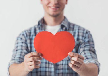 Yakışıklı olan rahat kıyafetler içinde büyük bir kırmızı kağıt tutan kalp ve gülümseyen, beyaz izole kırpılmış