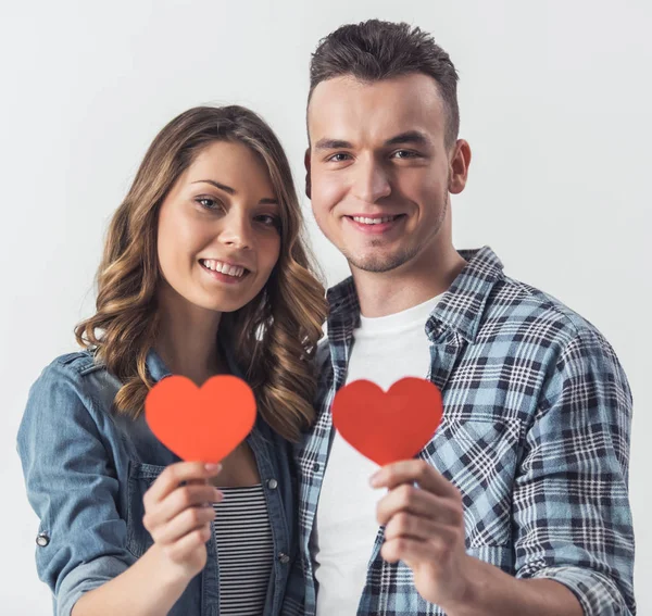 カジュアルな服で美しい若いカップルが つ赤い紙のハート カメラ目線と笑顔 白で隔離を保持しています — ストック写真