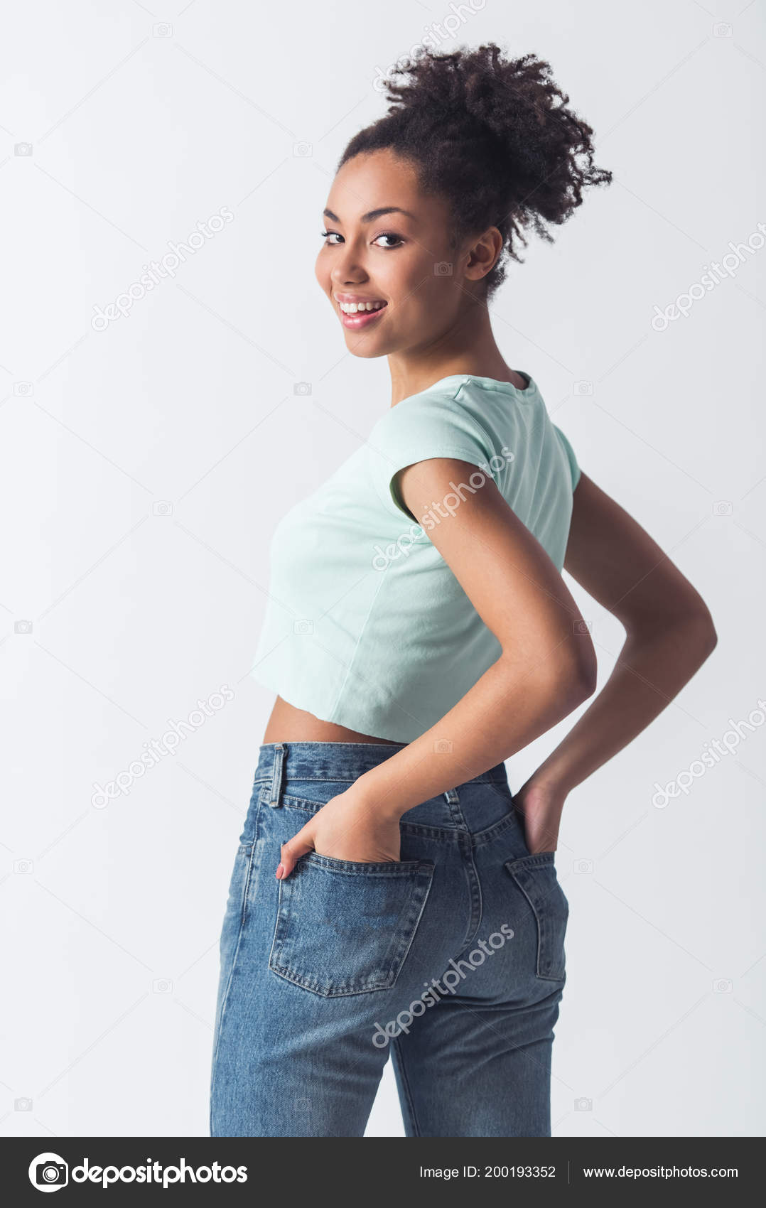 カジュアルな服で陽気なアフリカ系アメリカ人女の子がポーズをとって彼女のジーンズの後ろポケットに手を繋いでカメラ目線や笑顔 白で隔離 ストック写真 C Georgerudy