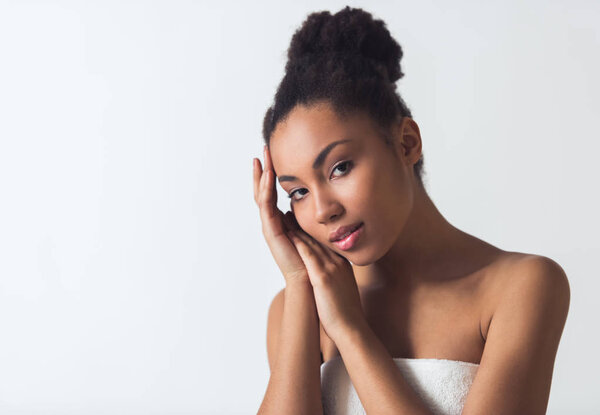 Beautiful Afro-American girl in towel is touching her face and looking at camera, isolated on white