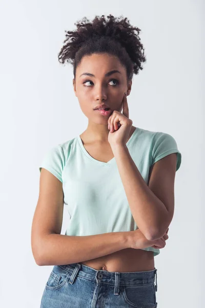 Beautiful Afro-American girl in casual clothes is touching her cheek, biting her lip, looking away and thinking, isolated on white