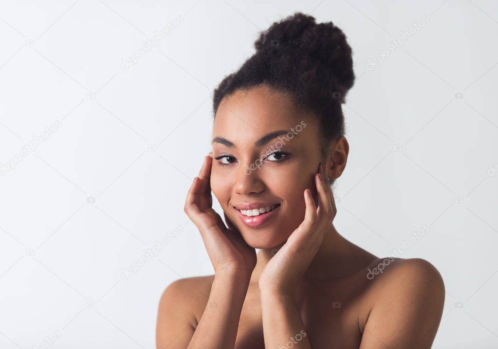 Beautiful Afro-American girl is touching her face, looking at camera and smiling, isolated on white