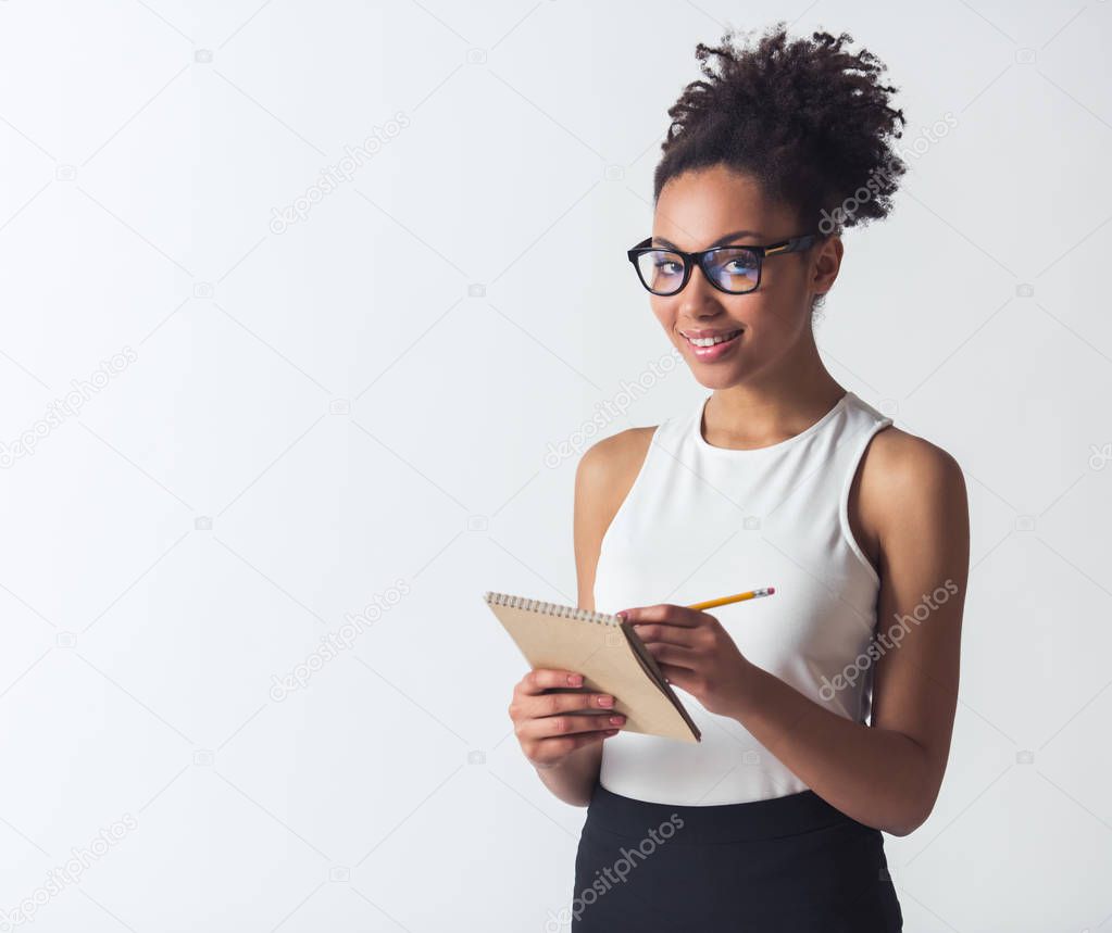 Beautiful Afro-American girl in eyeglasses is holding a notepad and a pencil, looking at camera and smiling, isolated on white