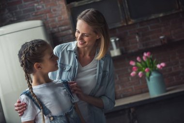 Anne ve kızı rahat kıyafetler içinde sarılma, birbirine bakarak ve evde mutfakta dururken gülümseyen