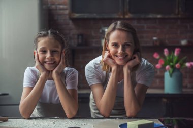 Güzel anne ve kızı önlük içinde mutfak masasında yaslanmış, kameraya bakıyor ve evde yemek yaparken gülümseyen
