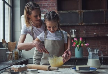Güzel anne ve kızı önlük içinde yumurta çırpma ve evde kurabiye yaparken gülümseyen