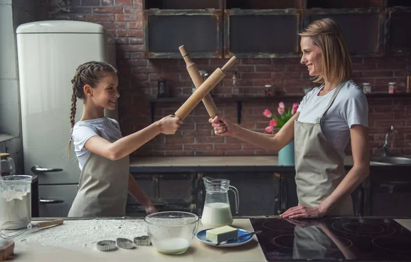 Beautiful mom and daughter in aprons are pretending to fight with rolling pins, looking at camera and smiling while cooking at home