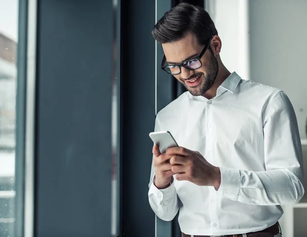 Hombre Negocios Guapo Gafas Está Utilizando Teléfono Inteligente Sonriendo Mientras —  Fotos de Stock