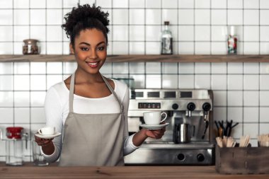 Güzel Afro Amerikan barista önlük bardak holding, kameraya bakıyor ve sayaç bar, dururken gülümseyen