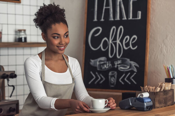 Beautiful Afro American barista in apron is holding a cup and smiling while standing at bar counter
