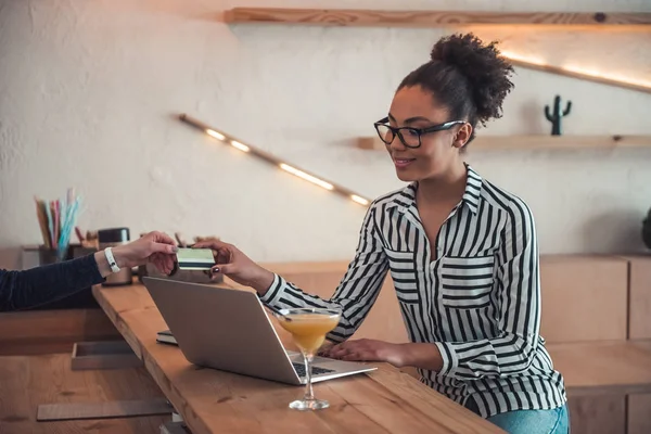 Beautiful Afro American girl in smart casual clothes and glasses is giving a credit card and smiling while working with a laptop in cafe