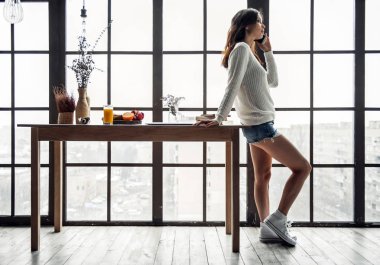 Güzel genç kız rahat kıyafetler içinde cep telefonu evde, ahşap masanın üzerine eğilerek tam uzunlukta konuşuyor