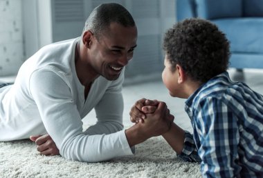Bilek güreşi ve halıya evde yalan gülümseyen mutlu Afro Amerikan baba ve oğul rahat kıyafetler içinde vardır