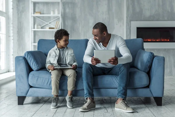 快乐的美国黑人父亲和儿子在休闲服装使用数码片和谈话 而坐在沙发上 爸爸在微笑 — 图库照片