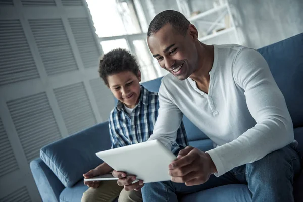 快乐的美国黑人父亲和儿子在休闲服装使用数码片和微笑 而坐在沙发上在家 — 图库照片