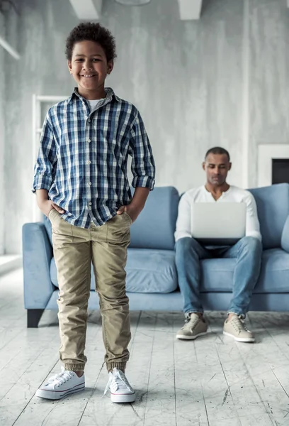 美国黑人男孩正在看着相机和微笑 而他的父亲正在使用笔记本电脑 坐在沙发上的背景 — 图库照片