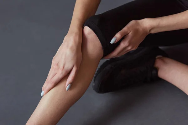 Cropped image of girl in sportswear touching her leg feeling pain while sitting on the floor on gray background