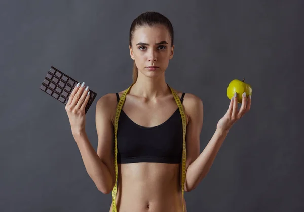 Beautiful girl in sportswear is holding an apple and chocolate bar and looking at camera, on gray background