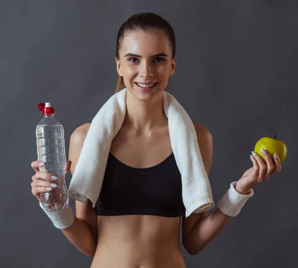 Beautiful girl in sportswear is holding an apple and a bottle of water, looking at camera and smiling, on gray background