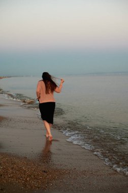 Gün batımında sahilde dalgaların yanında yürüyen bir kadın. 