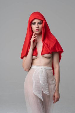 Stüdyoda poz veren genç ve güzel bir kız, beyaz şeffaf giysiler ve kırmızı bir eşarpla duruyor.
