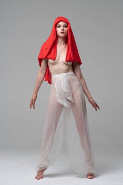 Stüdyoda poz veren genç ve güzel bir kız, beyaz şeffaf giysiler ve kırmızı bir eşarpla duruyor.