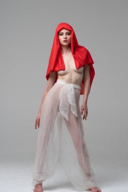 Stüdyoda poz veren genç ve güzel bir kız, beyaz şeffaf giysiler ve kırmızı bir eşarpla duruyor.