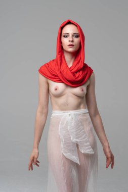 Stüdyoda poz veren genç ve güzel bir kız, beyaz şeffaf giysiler ve kırmızı bir eşarpla duruyor.